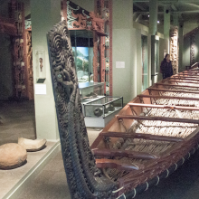 A large canoe with wooden rows and red detailing. The canoe sits amidst a museum with items from the collection surrounding it.