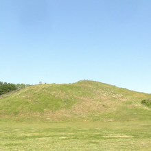 A large, earthen mound covered in grass set against a blue sky. The mound has stairs with people using them. 
