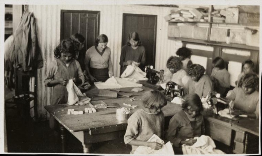 Black and white photo of 11 girls and one teacher some sitting some standing at tables with needles, fabric, and sewing machines. Girls are dark-skinned. Teacher is white.