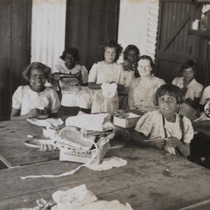 Black and white photo of 10 girls and one teacher seated at tables with needles, fabric, and sewing machines.