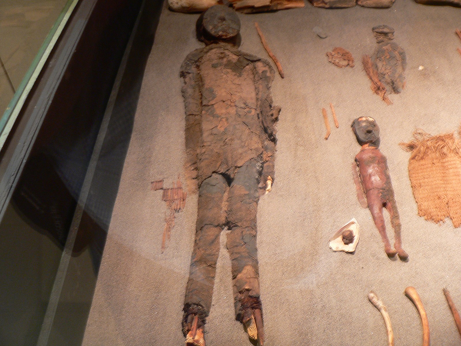 The mummified remains of an adult person and a child lay in a glass container for observation. The adult has a clay mask and black hair attached to their skull. The child lays next to the adult, with red paint on their body and a clay mask. 