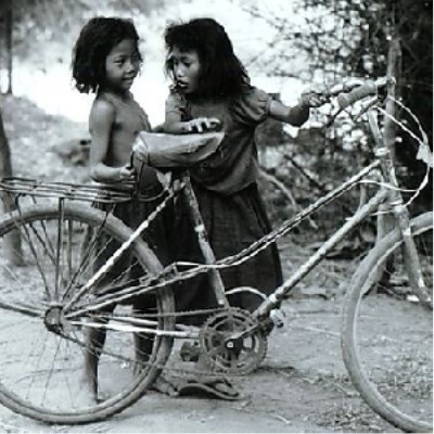 The image is titled on the site as "Children contend with a too-big bicycle, Pre Umbel" taken in 1991.  It is a black and white photograph showing too girls holding up a bicycle, unable to climb onto it.