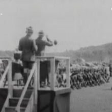 Black and white still of boy scouts marching past a reviewing stand. 